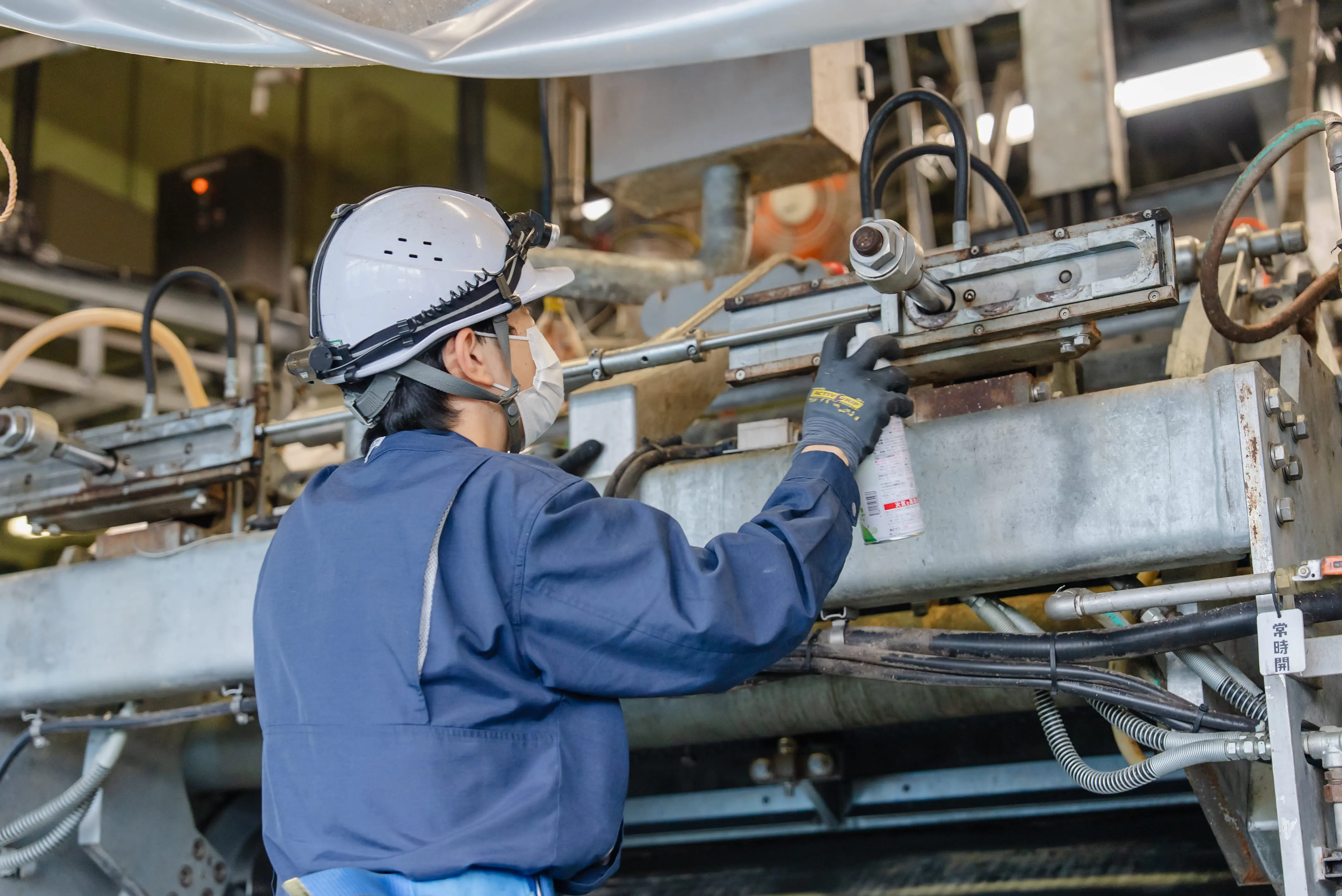 作業員が、配管や金属部品が並ぶ産業機械に手を伸ばし、機器の状態を確認している工場内作業風景
